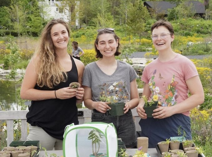 three people hold milkweed seedlings in pots and pose in a garden